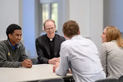 Father Robert A. Dowd, C.S.C., speaking with a group of three students seated at a table.